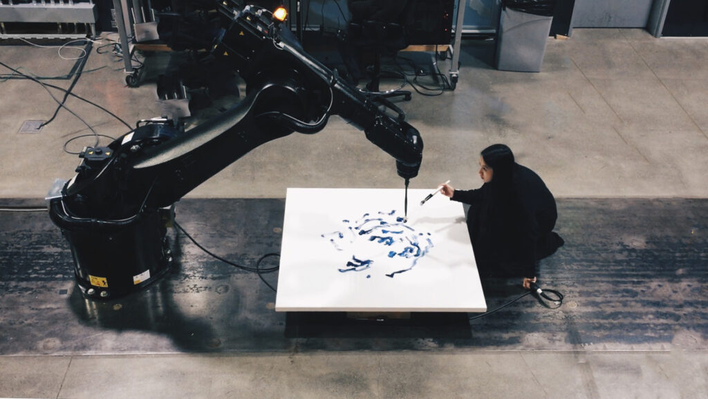 Photographie de Sougwen Chung agenouillée devant une toile posée au sol. En face d'elle, un bras robotique géant de sa conception tient un pinceau et peint en duo avec elle.