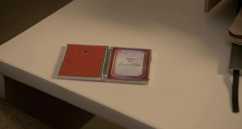 Extrait du film Her réalisé par Spike Jonze. Sur une table est posé ce qui ressemble à un petit carnet métallique. Il s'agit de la mini tablette pliable faisant office de smartphone dans le film. Sur la partie liseuse est écrit "Hello, I'm Samantha" soit "Bonjour, je suis Samantha" en français. Le ton de l'image est doux, beige, orange et rouge.